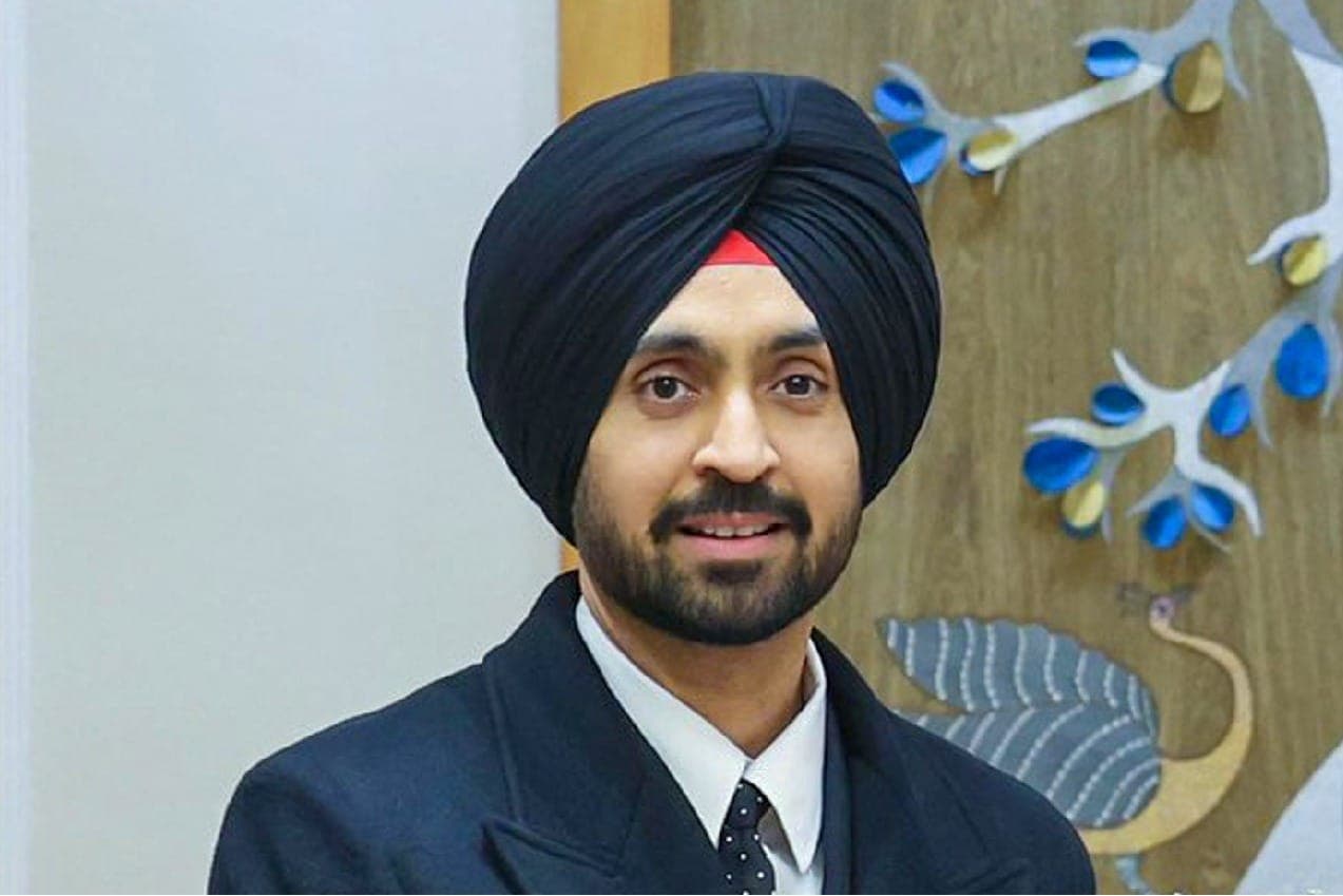 Diljit Dosanjh wearing a black turban, black suit and tie, smiling during an indoor appearance.